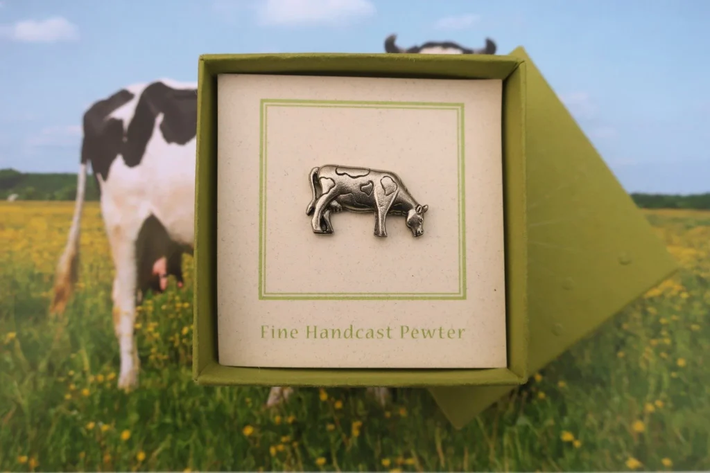 Pewter cow figurine in green box with photo of real cow in yellow flower field.