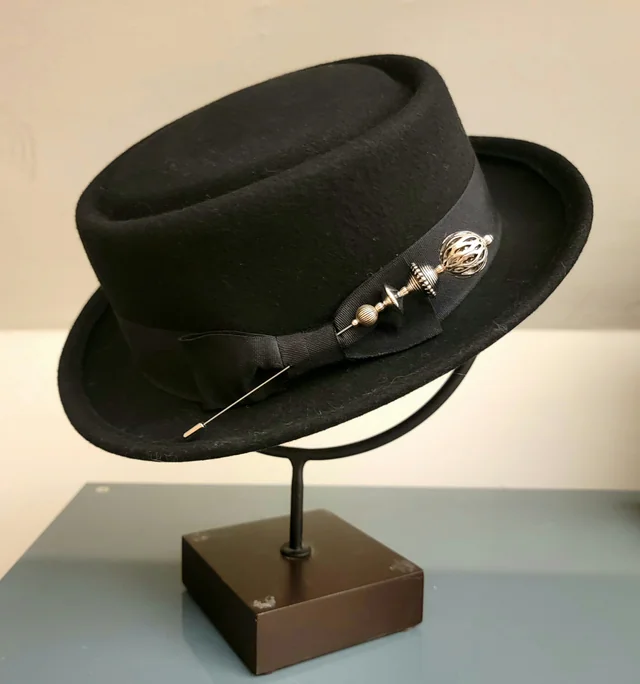 Black felt hat on a stand, decorated with a slim metal hatpin and silver beads on the hatband.